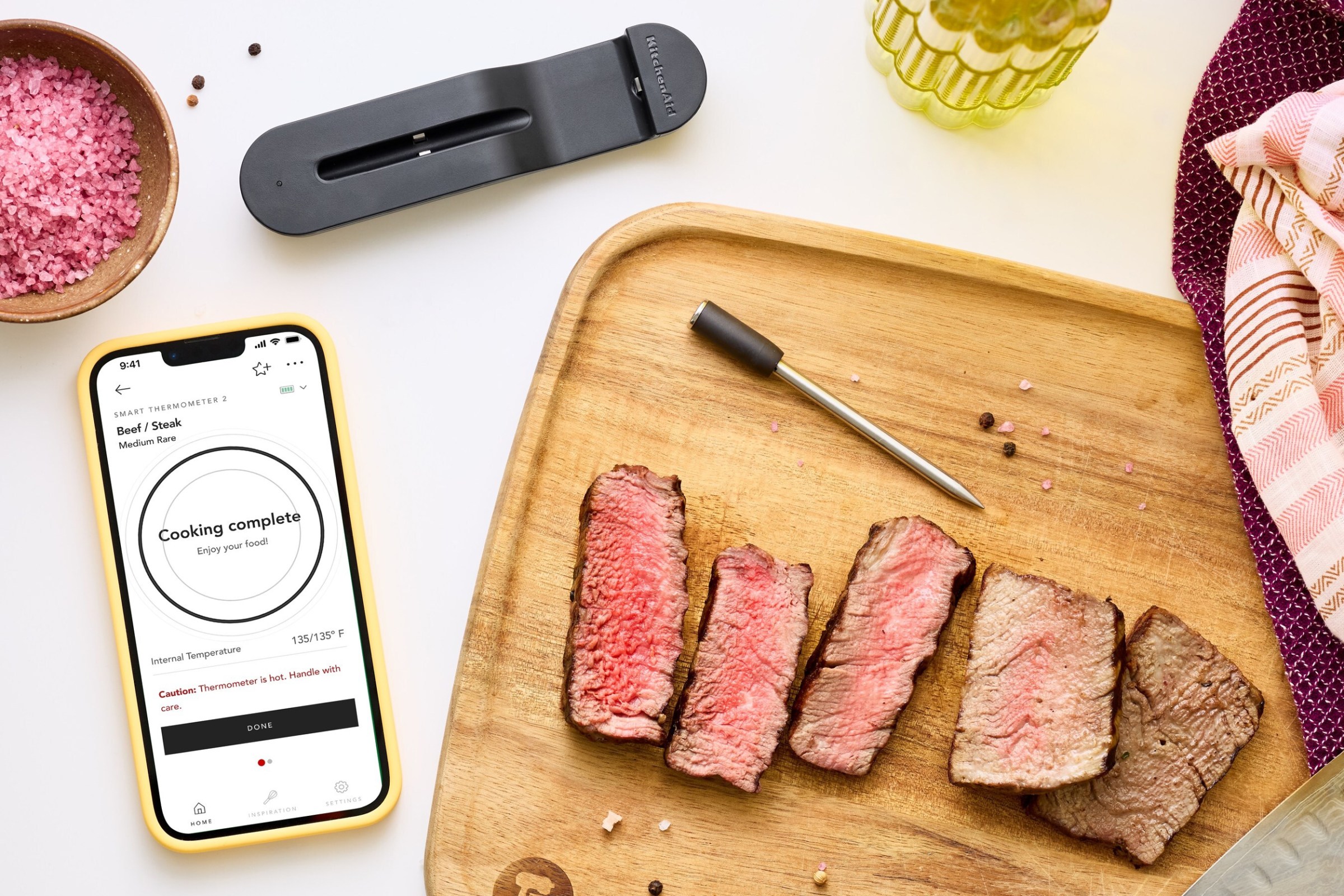 Several pieces of cooked steak on a wooden cutting board next to KitchenAid’s wireless Smart Thermometer and a smartphone.